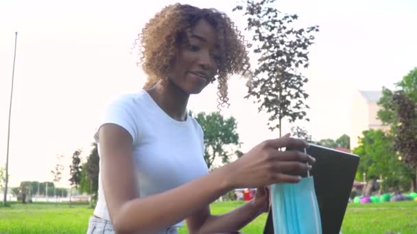 Giovane donna afroamericana che usa il computer portatile nel parco durante la pandemia del Covid 19. Maschera monouso medico appeso in mostra — Video Stock