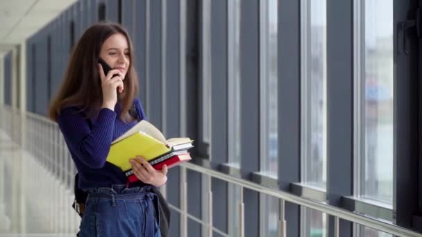 Kitapları olan bir kız öğrenci üniversitede telefonla konuşuyor. Sınavdan sonra mutlu öğrenci. Yeni modern tam işlevsel eğitim tesisi. Çevrimiçi eğitim kavramı — Stok video