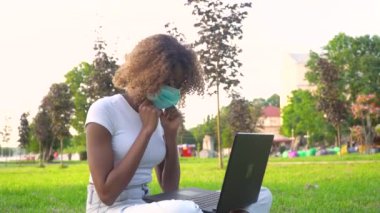 Genç Afrikalı Amerikalı kız öğrenci koruyucu maske takıyor ve parkta dizüstü bilgisayar üzerinde çalışıyor.