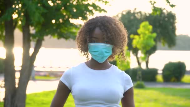 Afroamerykanka w masce medycznej do zapobiegania koronawirusowi i patrzenia w kamerę w parku. Ochrona przed Covid-19 — Wideo stockowe