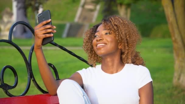 Mujer africana feliz hace selfie mientras está sentado en un banco en el parque — Vídeos de Stock