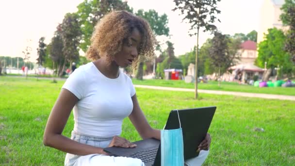 Jovem afro-americana convocando laptop conversa com professor online estudando no parque da cidade durante a pandemia de 19 anos. Máscara descartável médica pendurada na exposição — Vídeo de Stock