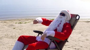 Güneş gözlüklü Noel Baba sahilde güneşlenirken kokteyl içer.