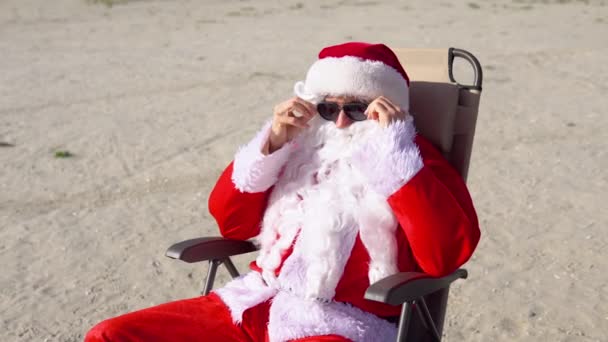 Güneş gözlüklü Noel Baba göl sahilinde güneşlenir. Seyahat ve tatil kavramı — Stok video