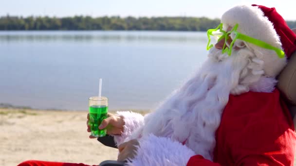 Père Noël dans des verres drôles boit un cocktail vert tout en étant couché sur une chaise longue sur la plage du lac. Concept de voyage et vacances — Video