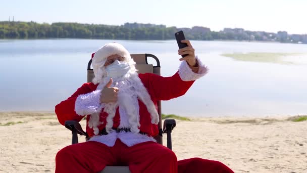 戴着白色防护面具的圣诞老人让自己躺在沙滩上的日光浴上。被隔离的圣诞老人 — 图库视频影像