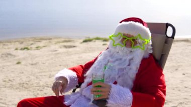 Komik gözlüklü Noel Baba sahilde güneşlenirken kokteyl içer.