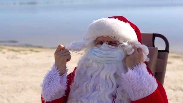 Le Père Noël enlève un masque médical couché sur une chaise longue sur la plage. Fin de l'épidémie — Video