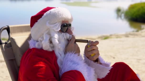 Güneş gözlüklü Noel Baba, tropik bir sahilde güneşlenirken puro içiyor. — Stok video