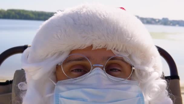 Papai Noel em uma máscara protetora branca deitada em uma espreguiçadeira na praia. O Pai Natal em quarentena. Movimento lento — Vídeo de Stock
