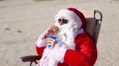 Güneş gözlüklü Noel Baba göl sahilinde güneşlenirken kokteyl içer. Noel Baba tropiklerde