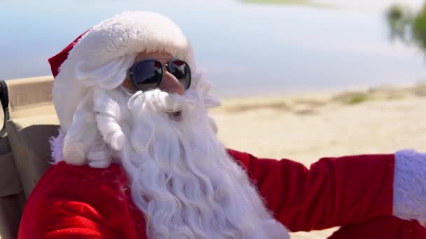 Babbo Natale in occhiali da sole fuma un sigaro cubano mentre si trova su un lettino su una spiaggia tropicale. Babbo Natale ai tropici — Video Stock
