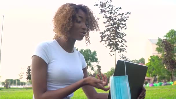 Joven mujer afroamericana llamando a la computadora portátil habla con el profesor en línea que estudia en el parque de la ciudad durante la pandemia covid 19. Máscara médica desechable colgando en la pantalla — Vídeos de Stock