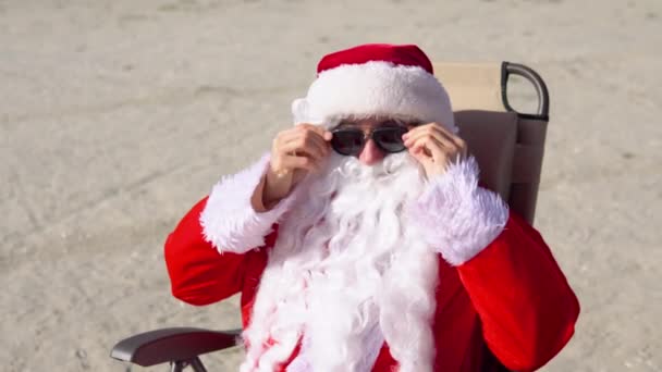 De kerstman doet een zonnebril op terwijl hij op een ligstoel op het strand ligt. Kerstman in de tropen — Stockvideo