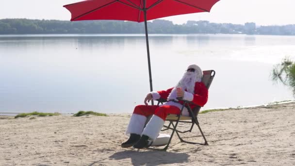 Babbo Natale in occhiali da sole beve un cocktail mentre sdraiato su un lettino sulla spiaggia del lago. Viaggio e concetto di vacanza — Video Stock