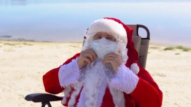 Beyaz koruyucu maskeli Noel Baba kumsalda güneşli bir yerde yatıyor. Noel Baba karantinada