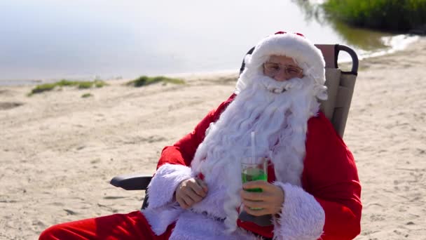 サンタクロースはビーチのサンラウンジャーに横たわっている間にカクテルを飲みます — ストック動画