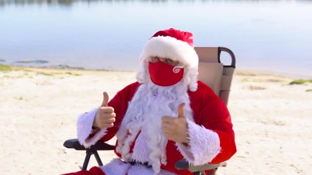 Babbo Natale con una maschera protettiva rossa sdraiato su un lettino sulla spiaggia. Babbo Natale in quarantena — Video Stock