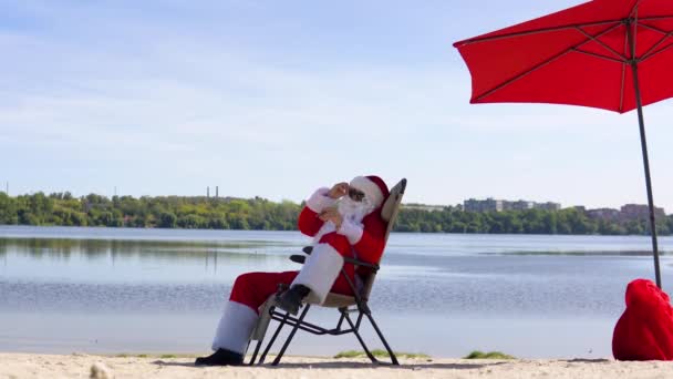 Güneş gözlüklü Noel Baba, göl kıyısında güneşlenirken puro içiyor. — Stok video