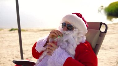 Güneş gözlüklü Noel Baba göl sahilinde güneşlenirken kokteyl içer. Seyahat ve tatil kavramı