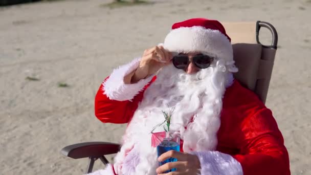 Le Père Noël en lunettes de soleil boit un cocktail tout en étant allongé sur une chaise longue sur la plage du lac. Père Noël sous les tropiques — Video