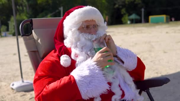 Le Père Noël boit un cocktail vert tout en étant allongé sur une chaise longue sur la plage — Video