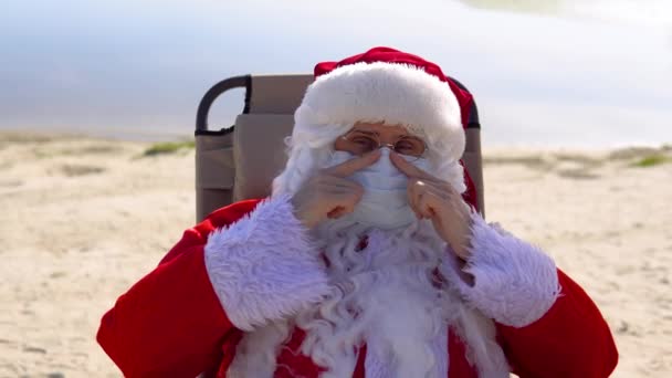 Santa Claus i en vit skyddsmask som ligger på en solstol på stranden. Jultomten i karantän — Stockvideo