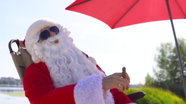 Père Noël en lunettes de soleil fume un cigare cubain alors qu'il est allongé sur une chaise longue sur une plage — Video