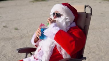 Güneş gözlüklü Noel Baba göl sahilinde güneşlenirken mavi bir kokteyl içer. Noel tatili.