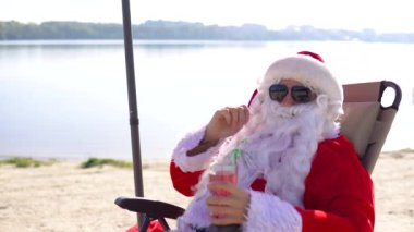 Güneş gözlüklü Noel Baba, göl sahilinde güneşlenirken kokteyl içer.