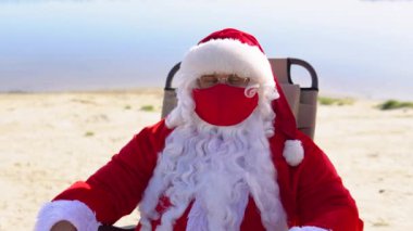 Kırmızı koruyucu maskeli Noel Baba kumsalda güneşli bir yerde yatıyor. Noel Baba karantinada