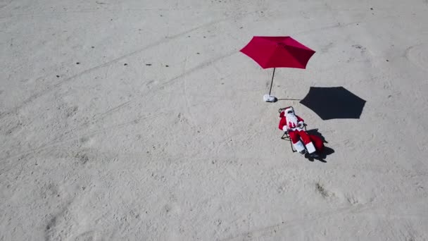 Babbo Natale è sdraiato su un lettino sulla spiaggia di sabbia. Vista dall'alto. La telecamera si sposta a destra. Babbo Natale ai tropici — Video Stock