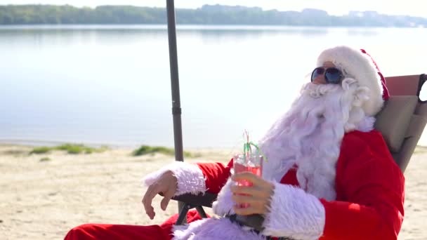 Le Père Noël en lunettes de soleil boit un cocktail allongé sur une chaise longue sur la plage du lac — Video