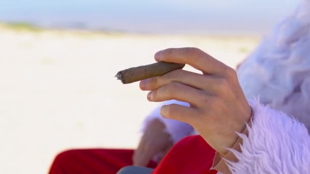 De kerstman in een zonnebril rookt een Cubaanse sigaar terwijl hij op een ligstoel op het strand ligt. Kerstman in de tropen — Stockvideo