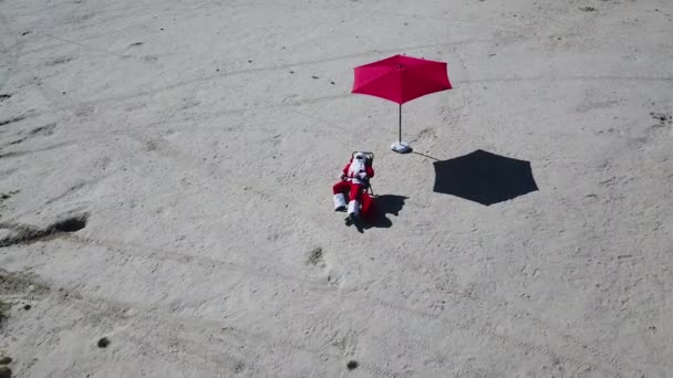 Le Père Noël se trouve sur une chaise longue sous un parapluie rouge sur une plage de sable. Vue de dessus. Père Noël sous les tropiques — Video