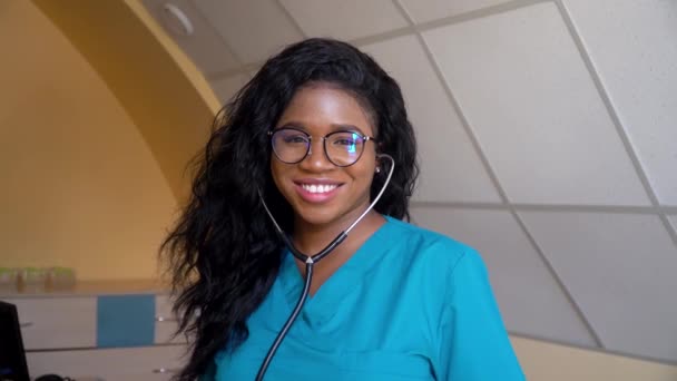 幸せなアフリカ系アメリカ人女性医師や看護師の専門家一般開業医は、医療事務所でカメラを見て聴診器でポーズをとります.医学、医療、人々の概念 — ストック動画
