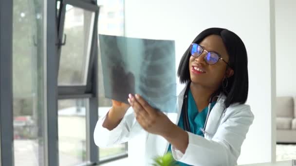 Afrikaanse vrouwelijke arts met bril kijken naar x-ray van de longen terwijl zitten aan tafel in de moderne kliniek — Stockvideo