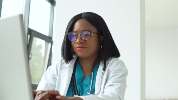 Jonge Afro-Amerikaanse studente in witte jas en stethoscoop over haar nek met behulp van laptop aan het bureau tijdens het werken op de universiteit. Onderwijsconcept — Stockvideo