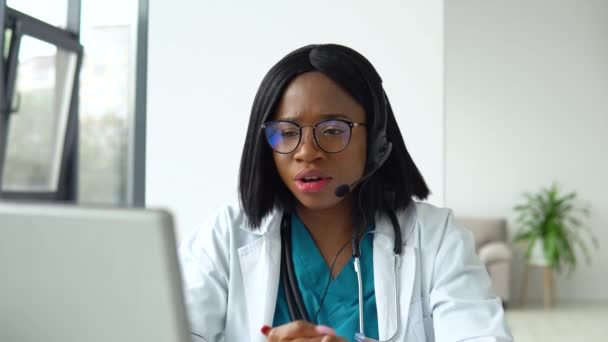 Beyaz önlüklü ve kulaklıklı profesyonel Afro-Amerikan kadın doktor laptop bilgisayarında konferans görüşmesi yapıyor, görüntülü sohbetteki hastaya danışmanlık yapıyor. — Stok video