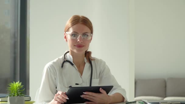 Női orvos fehér köpenyben és védőszemüvegben tablettával, kamerába néz, miközben orvosi rendelőben dolgozik — Stock videók