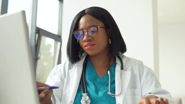 Joven estudiante afroamericana con bata blanca y estetoscopio sobre su cuello usando un portátil en el escritorio mientras trabajaba en la universidad. Concepto educativo — Vídeos de Stock