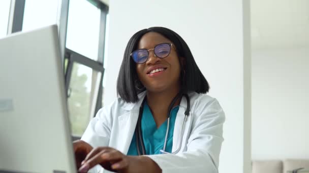 Szczęśliwa młoda, afrykańska, amerykańska lekarka z laptopem patrzącym w kamerę. Uśmiechnięta lekarka pozująca w gabinecie szpitalnym — Wideo stockowe