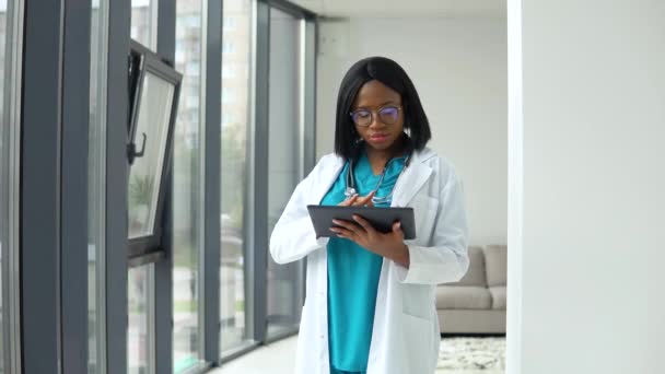 Afroamerikanische Ärztin in weißem Mantel und Stethoskop über dem Hals mit Tablette am Schreibtisch bei der Arbeit in der Arztpraxis — Stockvideo