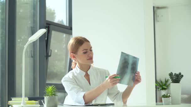 Женщина-врач смотрит на рентген легких, сидя за столом в современной клинике — стоковое видео