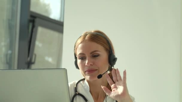 穿着白色医疗外套和耳机的专业女医生在笔记本电脑上进行电话会议，在视频聊天中在线咨询远程病人 — 图库视频影像