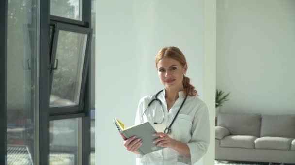 Jovem médica caucasiana feliz usando casaco médico branco e estetoscópio com bloco de notas olhando para a câmera. Médica sorridente posando no consultório do hospital — Vídeo de Stock