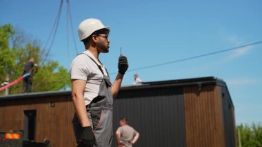 Güvenlik kaskı ve eldiven takan inşaat işçisi telsiz kullanıyor, prefabrik ev modülleriyle inşaat sahasında operasyonlar düzenliyor.