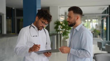 Laboratuvar önlüğü ve steteskop giyen profesyonel bir tıp uzmanı hastaların endişelerini ve semptomlarını dikkatle dinliyor ve modern hastane ortamındaki panolardaki bilgileri dikkatlice belgeliyor.