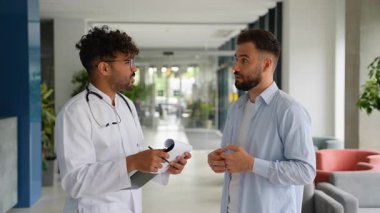 Hasta, doktor semptomlarını, boyun ve dirsek gibi farklı vücut parçalarını gösterirken, doktor hastane koridorundaki panoya not alıyor.