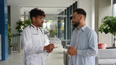 Hasta ile ilgilenirken danışmanlık detaylarını belgeleyen profesyonel erkek doktor, çağdaş tıbbi tesiste kendine güvenen bir tokalaşmayla sonuçlanıyor.